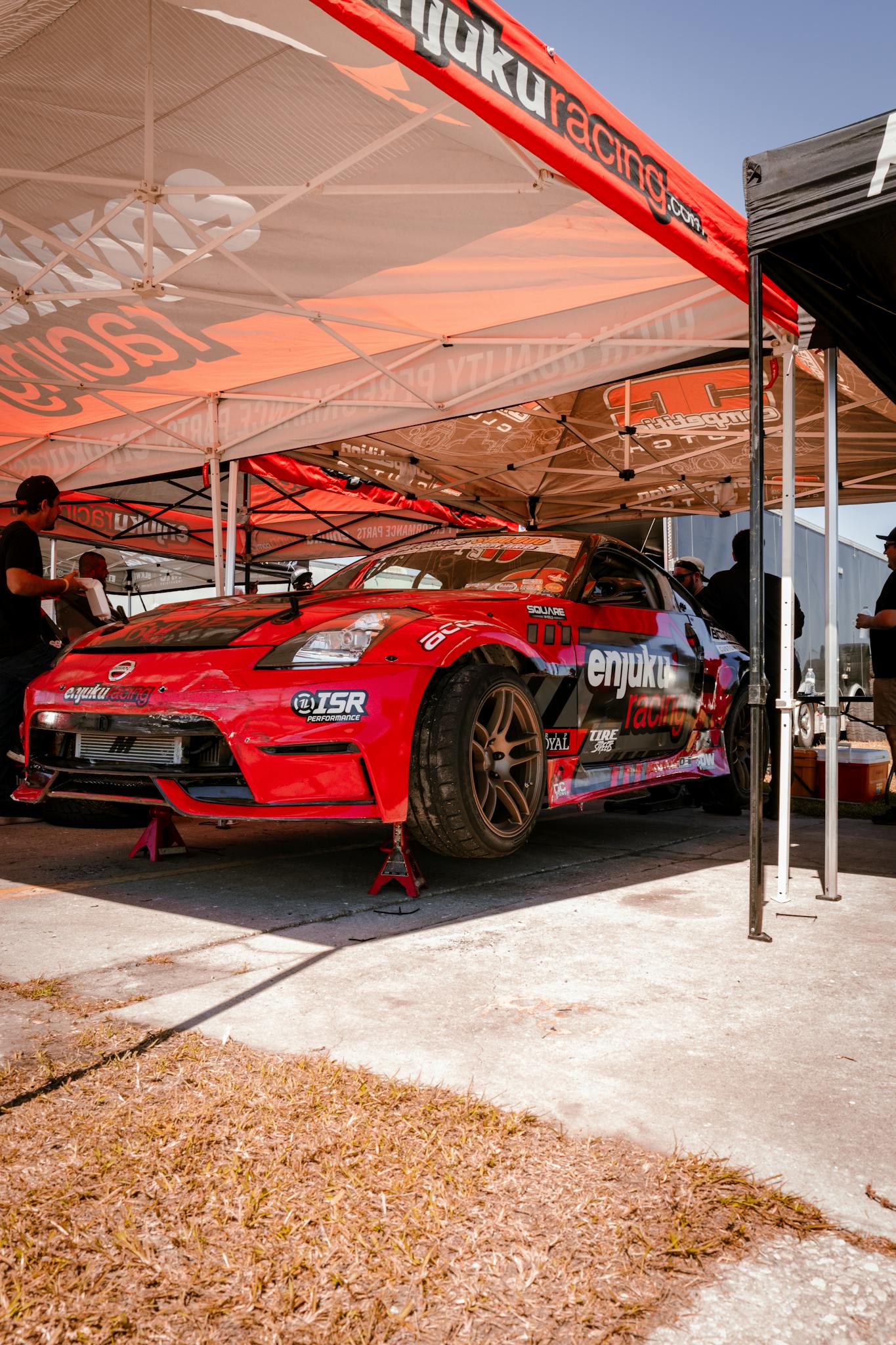 A red Nissan 350Z race car on jack stands at a motorsport event under a tent.